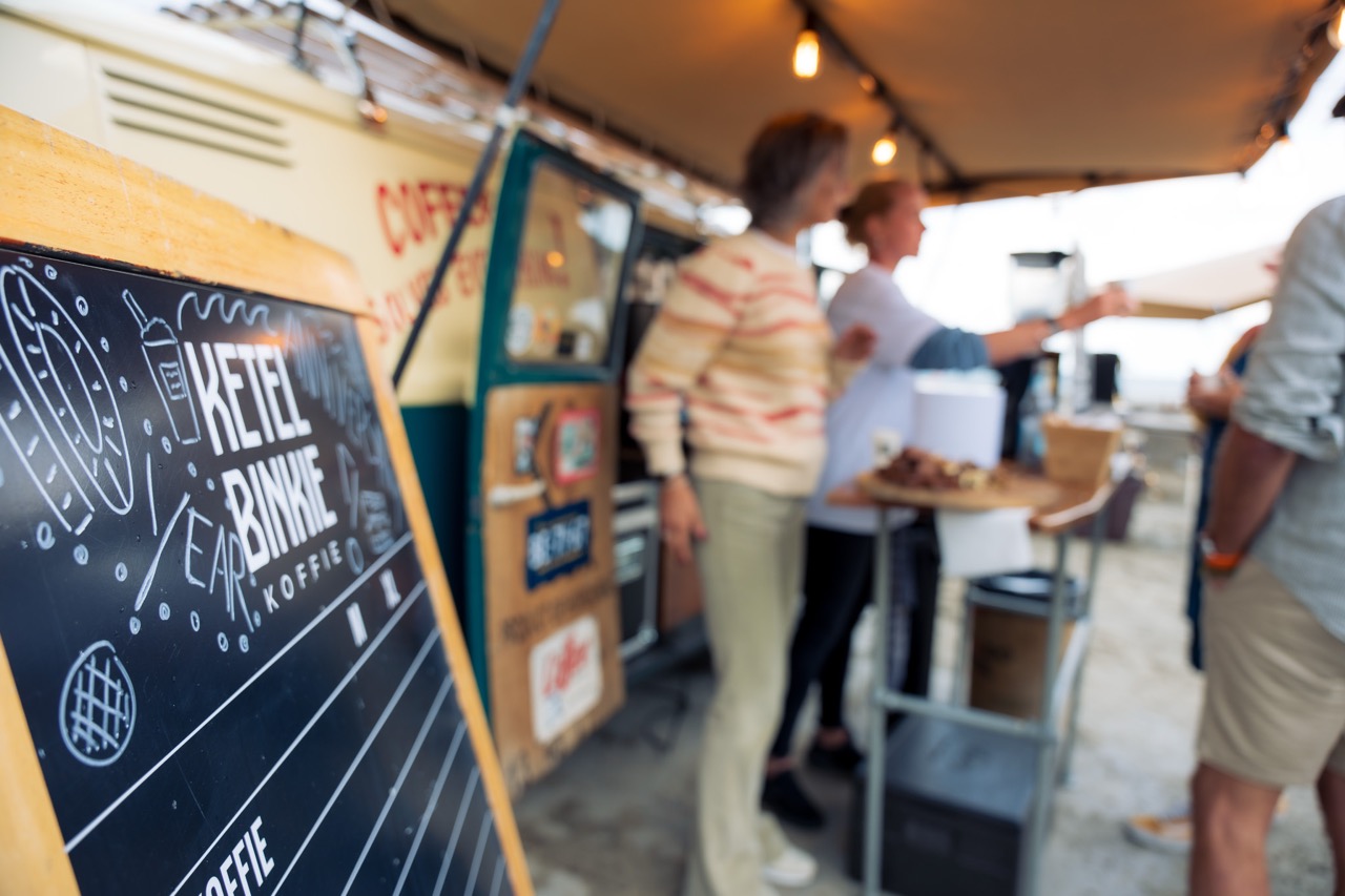 Krijtbord met tekst “KETELBINKIE KOFFIE” op terras, mensen drinken koffie onder luifel op het strand.
