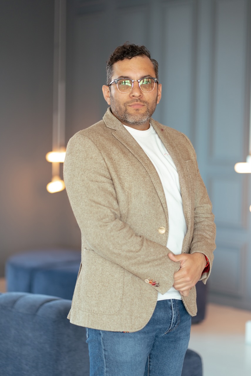 Man met bril en baard, draagt beige blazer en witte shirt, staand in moderne kantoorruimte.