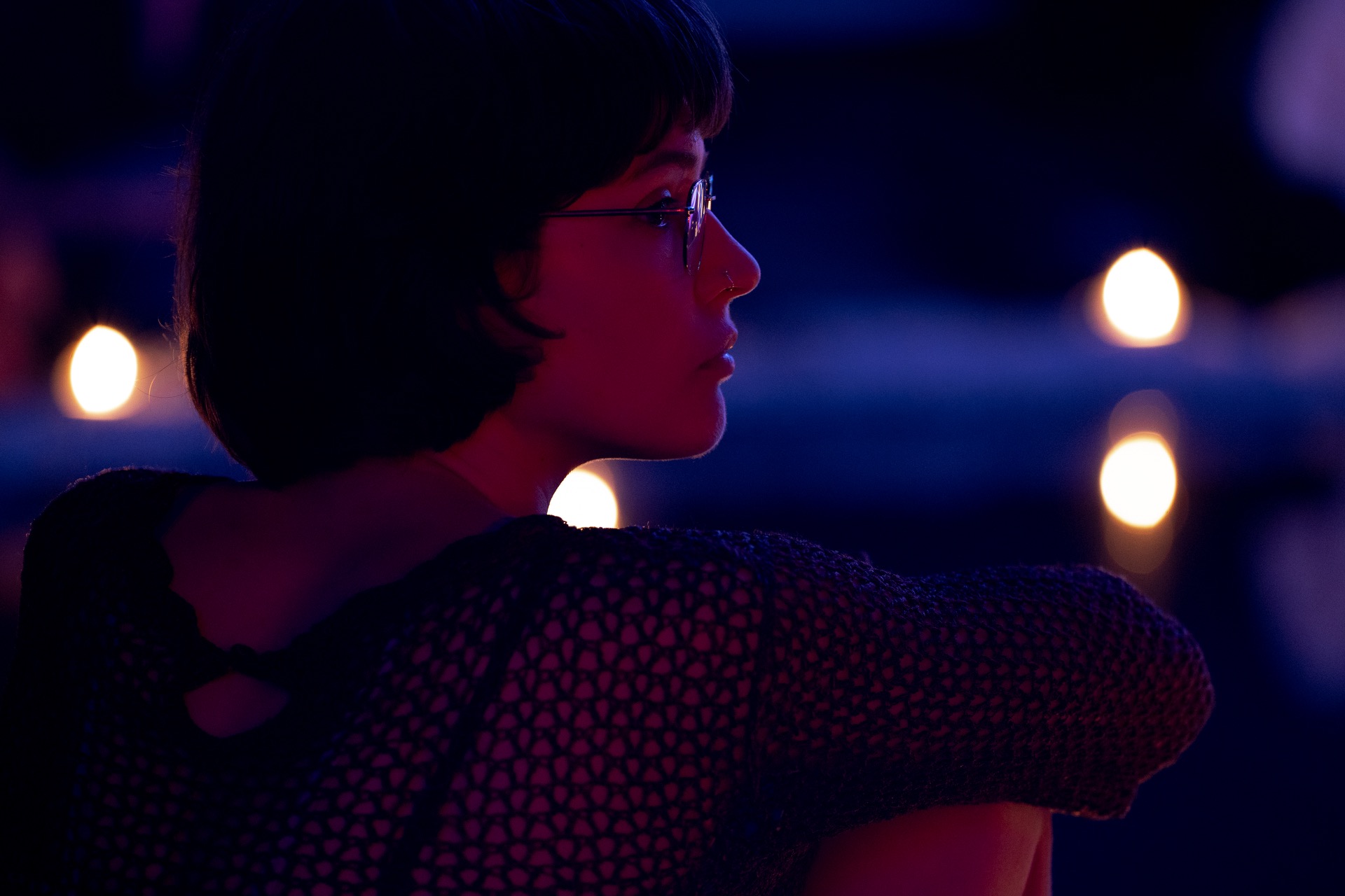 Portret van een vrouw in profiel met bril, belicht door paarse neonlichten en onscherpe bokeh.