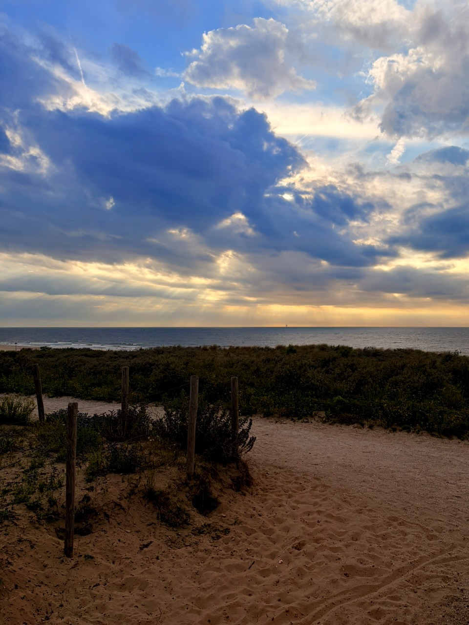 Zandstrand met duinen, lage struiken en houten palen, met zee en dramatische wolkenlucht bij zonsondergang.